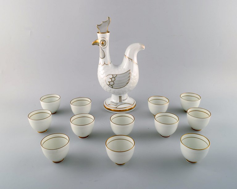 Bing & Grondahl/ B&G, Denmark. Jug of porcelain in the form of rooster, stopper 
with rooster comb. 12 accompanying porcelain mugs with gold-painted details.