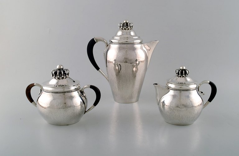 Sjældent Georg Jensen kaffeservice i sterlingsølv med hanke af ibenholt. 
Kaffekande, sukkerskål og flødekande. Låg udformet som kongekrone. Dateret 
1915-30.
